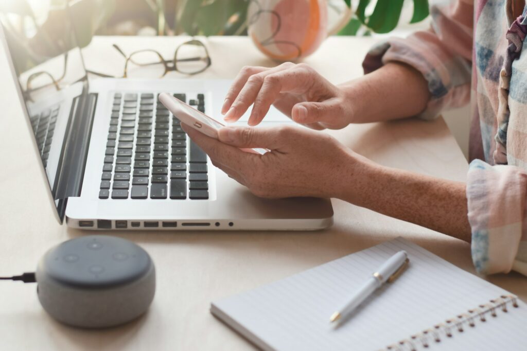 Woman sitting at desk with laptop computer and virtual assistant smart speaker using mobile phone